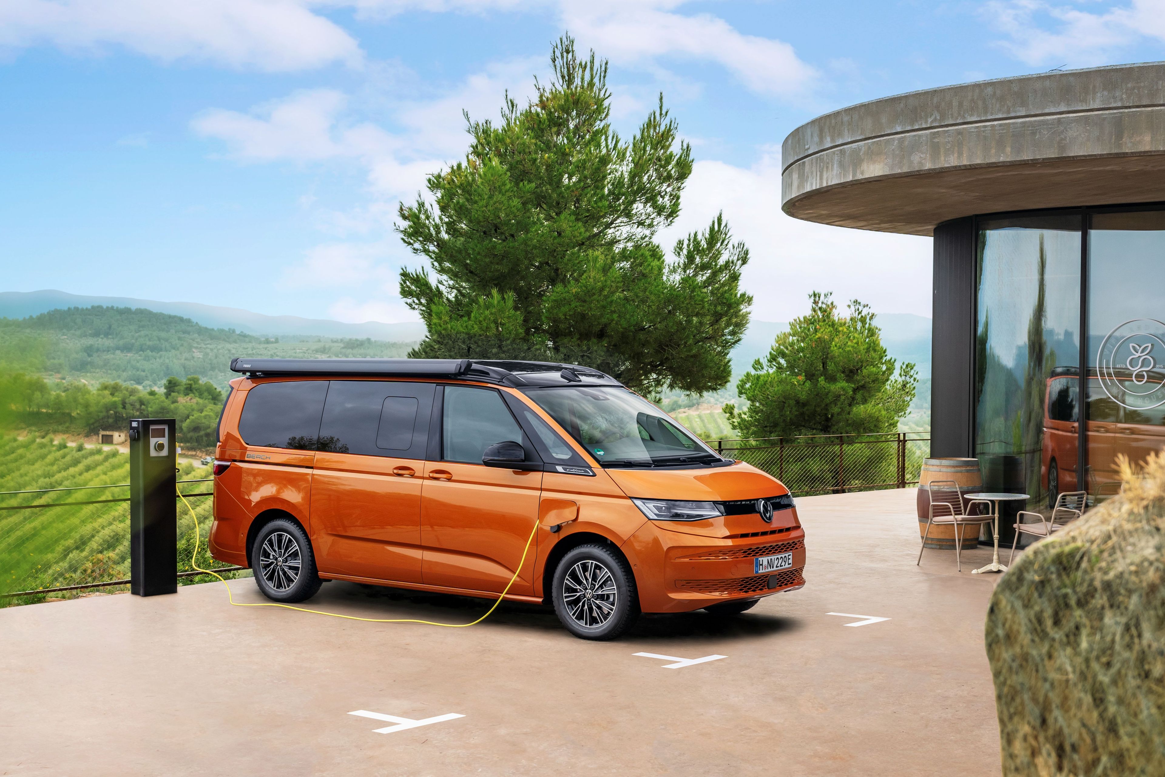 Oranžen kombi je parkiran ob sodobni zgradbi na polnilni postaji za električna vozila, obdan z zelenimi polji in drevesi. Zgradba ima steklene stene in zunanjo teraso s stoli in mizami. V ozadju se razprostirajo griči pod modrim nebom z nekaj oblaki.