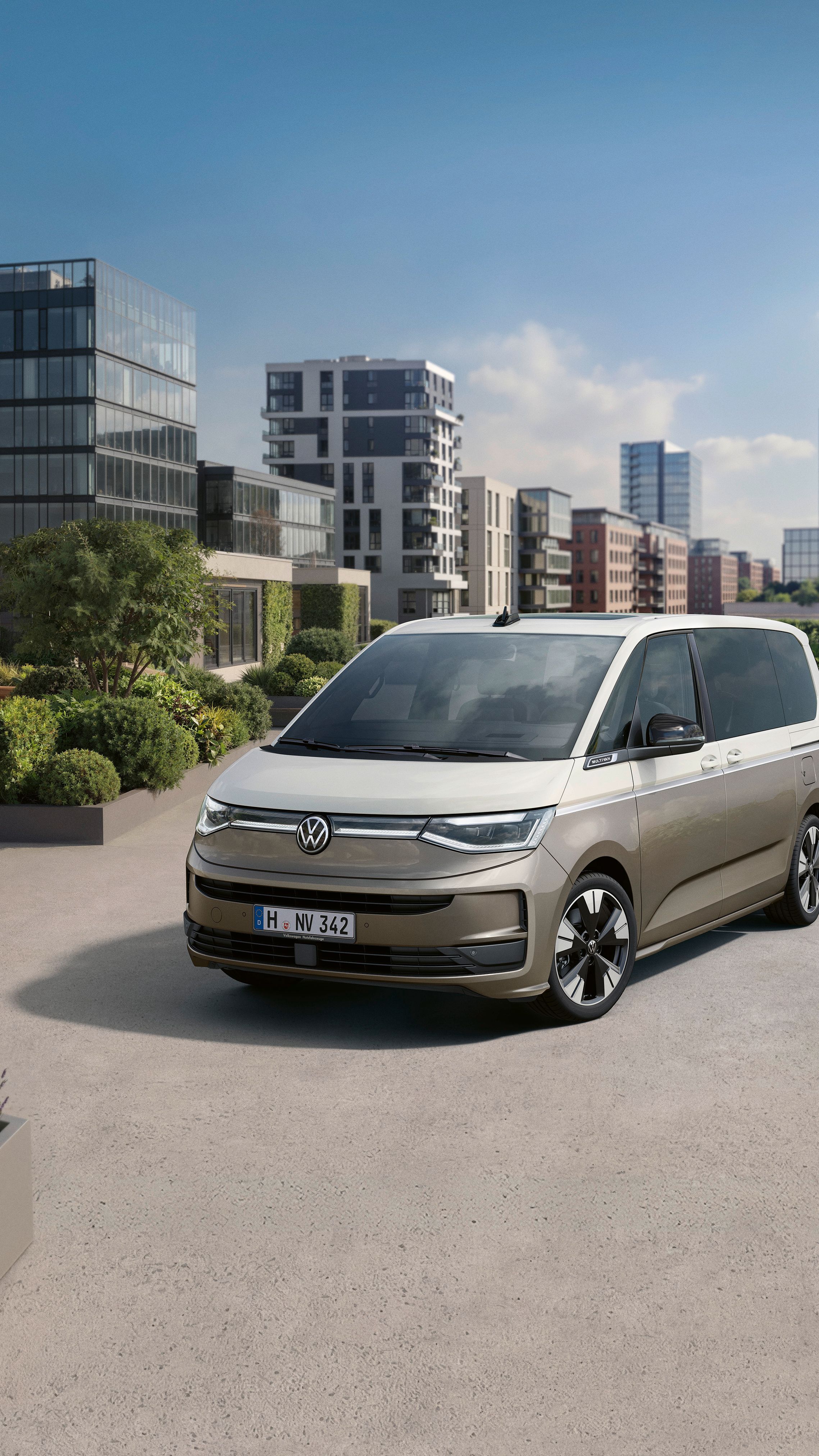 Sodobna srebrno-bež enoprostorec Volkswagen Multivan stoji na prostorni terasi z urejenimi zelenimi gredami. V ozadju so moderna poslovna in stanovanjska poslopja ter nebotičniki ob vodi pod jasnim modrim nebom. Atmosfera je urbana, mirna in sončna.