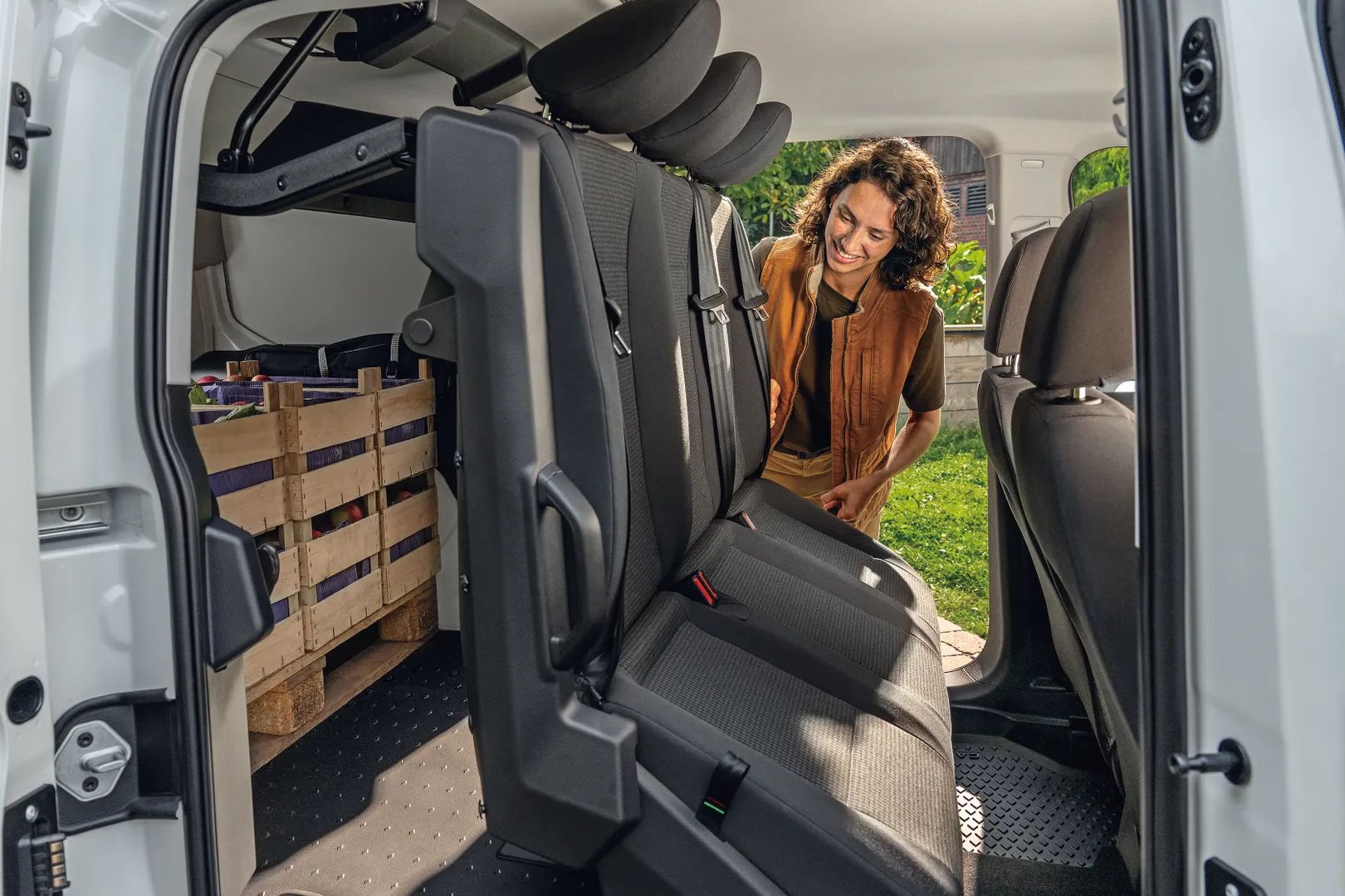 Oseba z zamegljenim obrazom stoji poleg odprtih vrat belega kombija. Notranjost kombija prikazuje zložen sedež in leseno škatlo za prenos tovora.