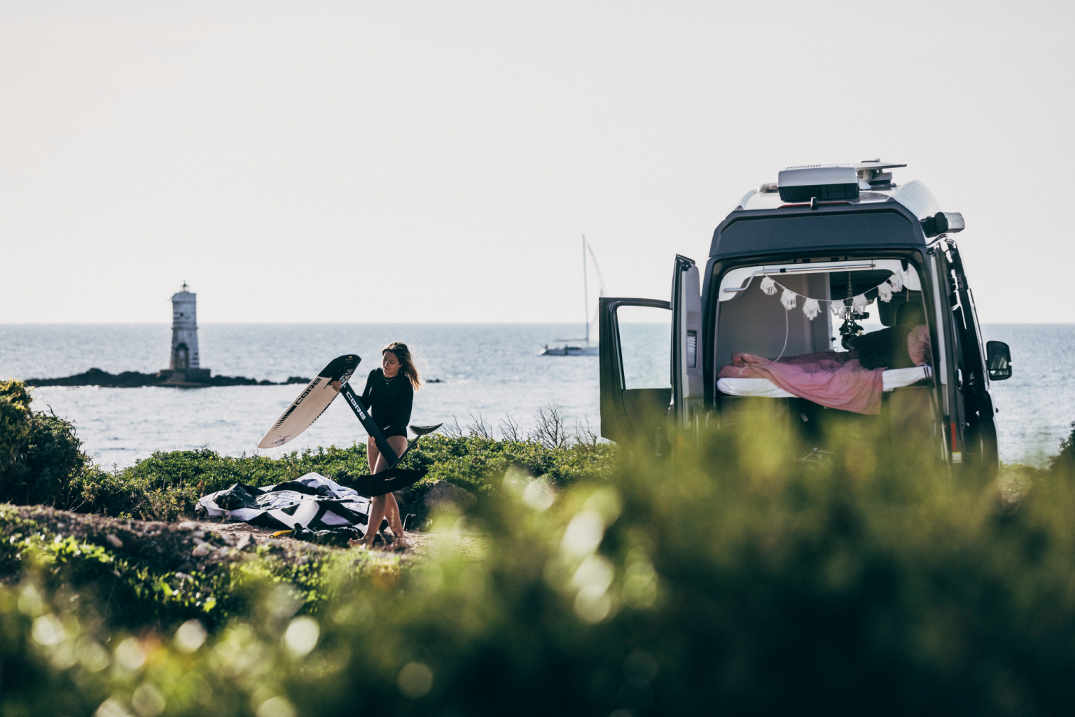 Ženska surfa pred kombijem z odprtimi vrati v obmorskem okolju. Ob obali je svetilnik, v ozadju pa jadrnica na morski gladini. Kombi ima znotraj okrašen prostor z lučkami.
