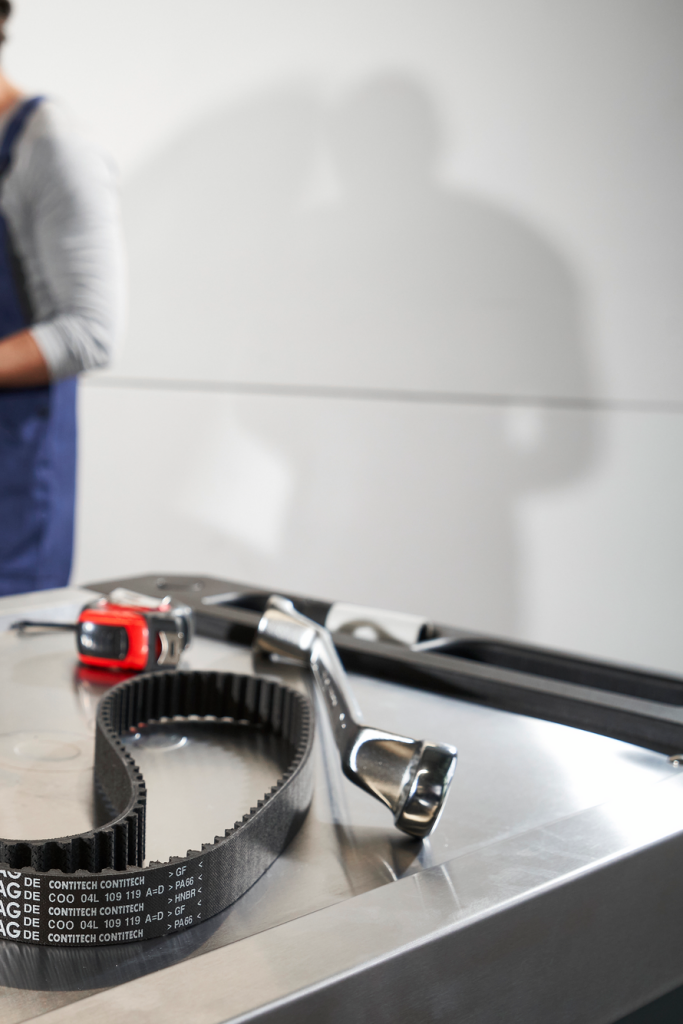 Na mizi v avtomehanični delavnici ležita zobati jermen in nekaj orodja, v ozadju je odprt pokrov avtomobila. Za avtomobilom stoji mehanik v predpasniku in na tablici preverja podatke ali zapisuje opombe. Scena prikazuje servis ali pregled vozila.