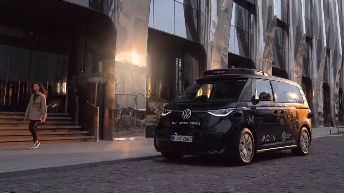 Ein selbstfahrender schwarzer Volkswagen-Van steht vor einem modernen Glasgebäude bei Sonnenuntergang. Eine Frau geht in der Nähe des Vans auf der Straße entlang.