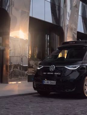 Ein selbstfahrender schwarzer Volkswagen-Van steht vor einem modernen Glasgebäude bei Sonnenuntergang. Eine Frau geht in der Nähe des Vans auf der Straße entlang.