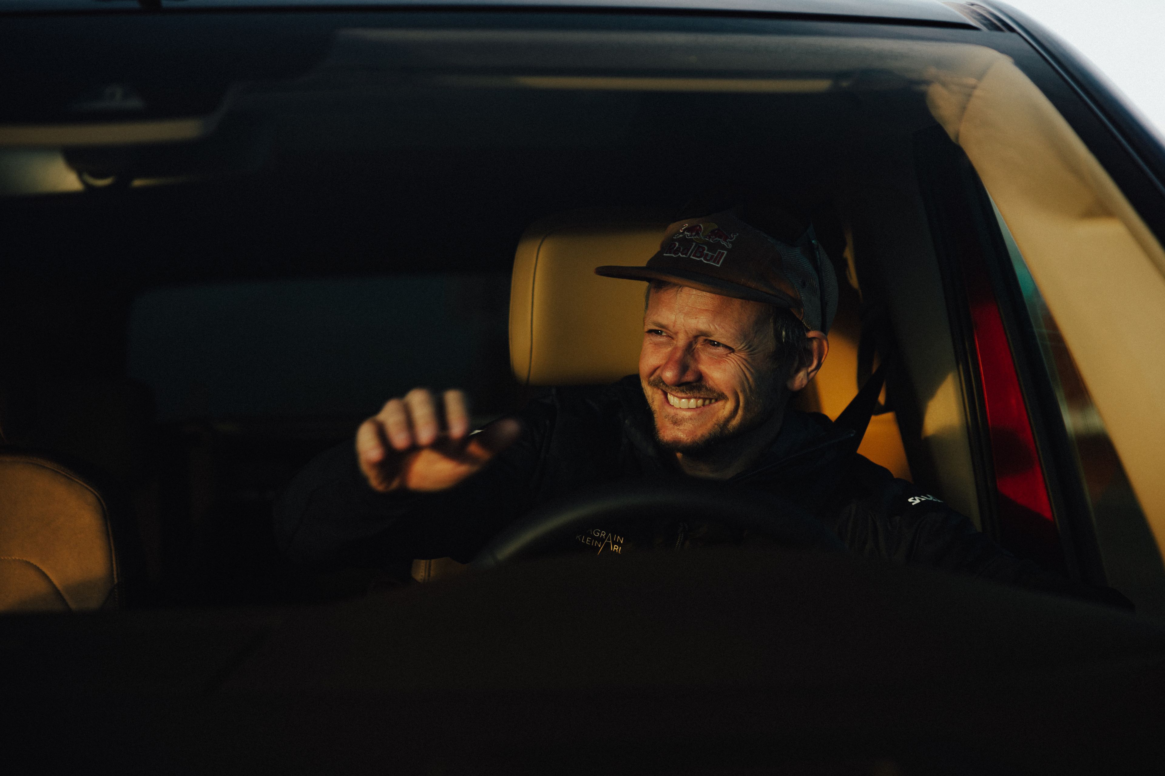 Oseba z zamegljenim obrazom sedi v avtu in vozi, del kabine avtomobila je osvetljen z naravno svetlobo. Roka je na volanu in vidijo se notranji deli avtomobila, kot sta naslonjalo sedeža ter nadzorna plošča.