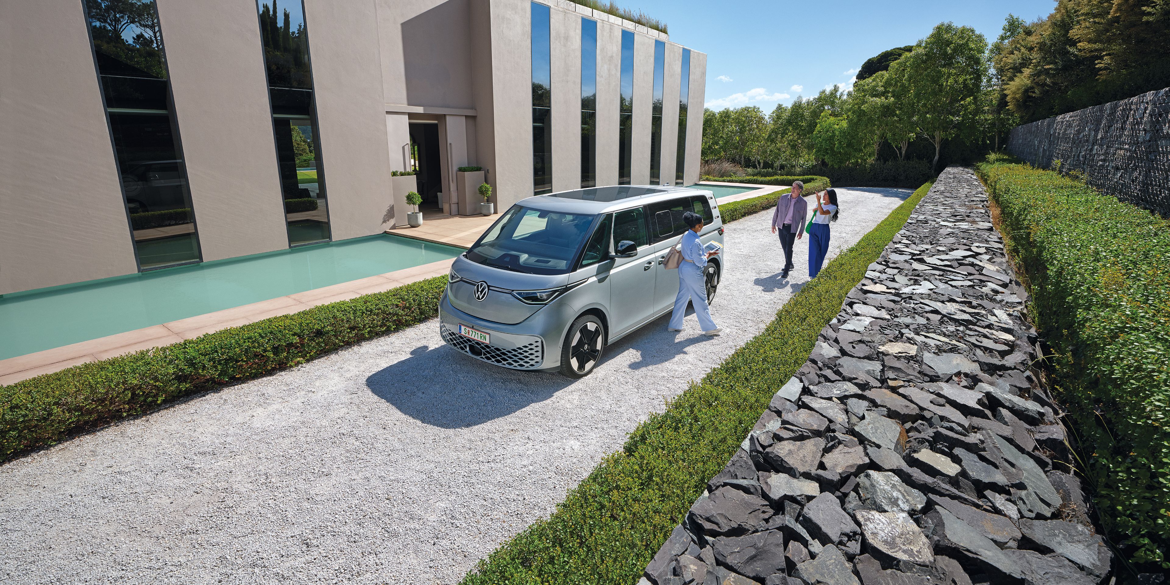 Ein silberner ID. Buzz parkt vor einem modernen Haus, ein Paar  geht auf das Auto zu