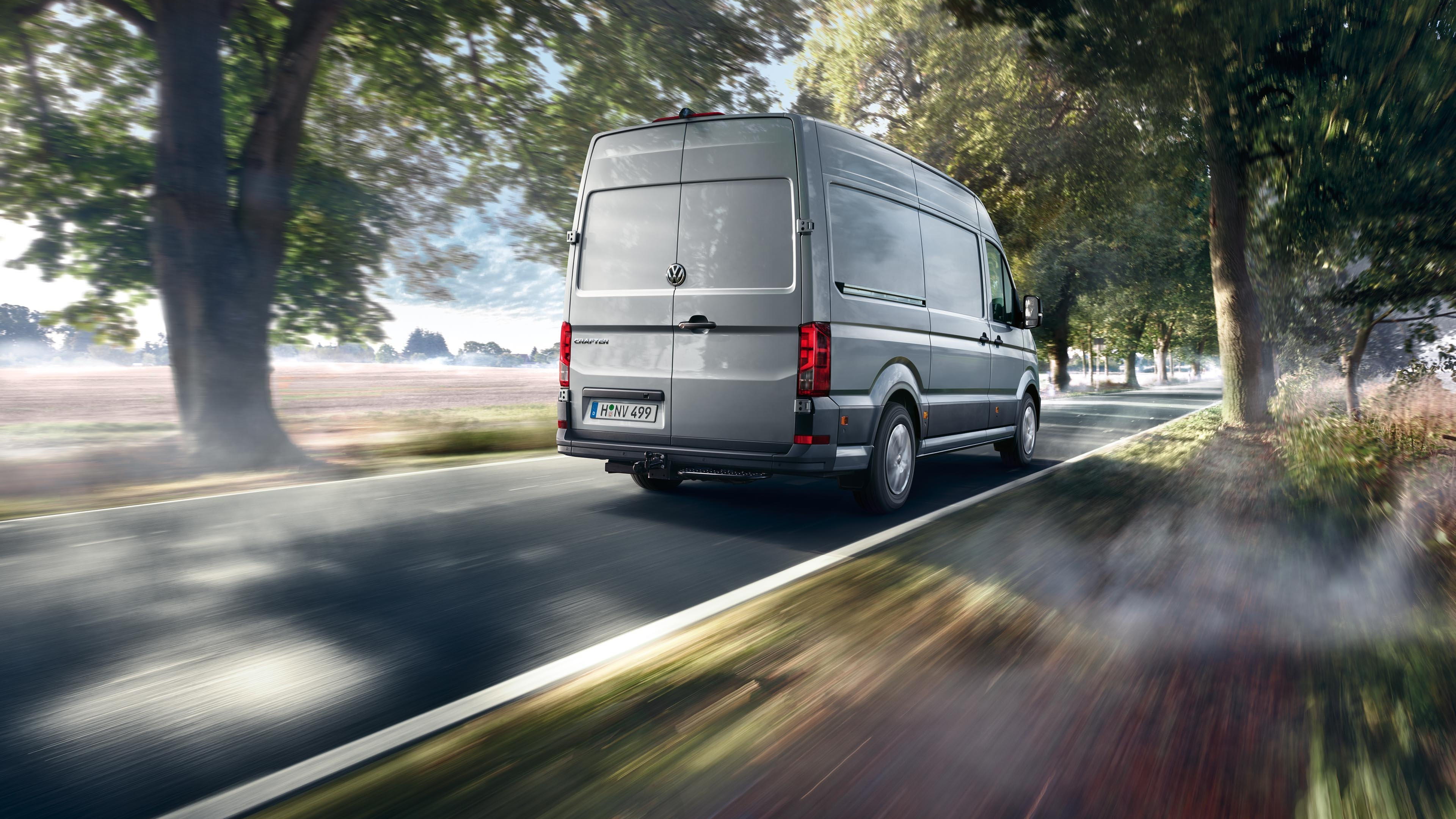 Siva dostavna kombi vozi po podeželski cesti, obdani z drevesi. Zgodaj zjutraj meglica prekriva del ceste. Pokrajina v ozadju je mirna in slikovita.