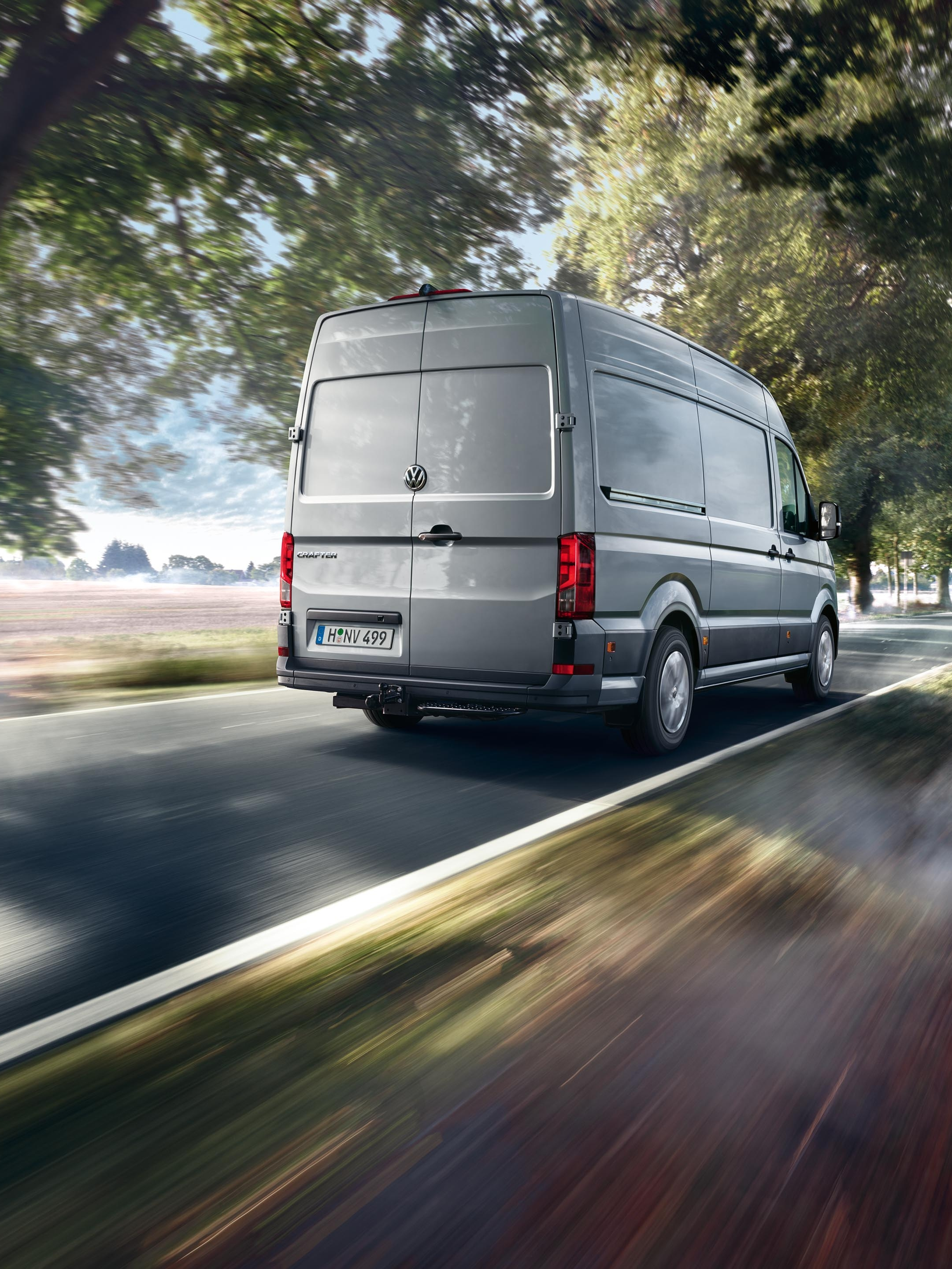 Siva dostavna kombi vozi po podeželski cesti, obdani z drevesi. Zgodaj zjutraj meglica prekriva del ceste. Pokrajina v ozadju je mirna in slikovita.