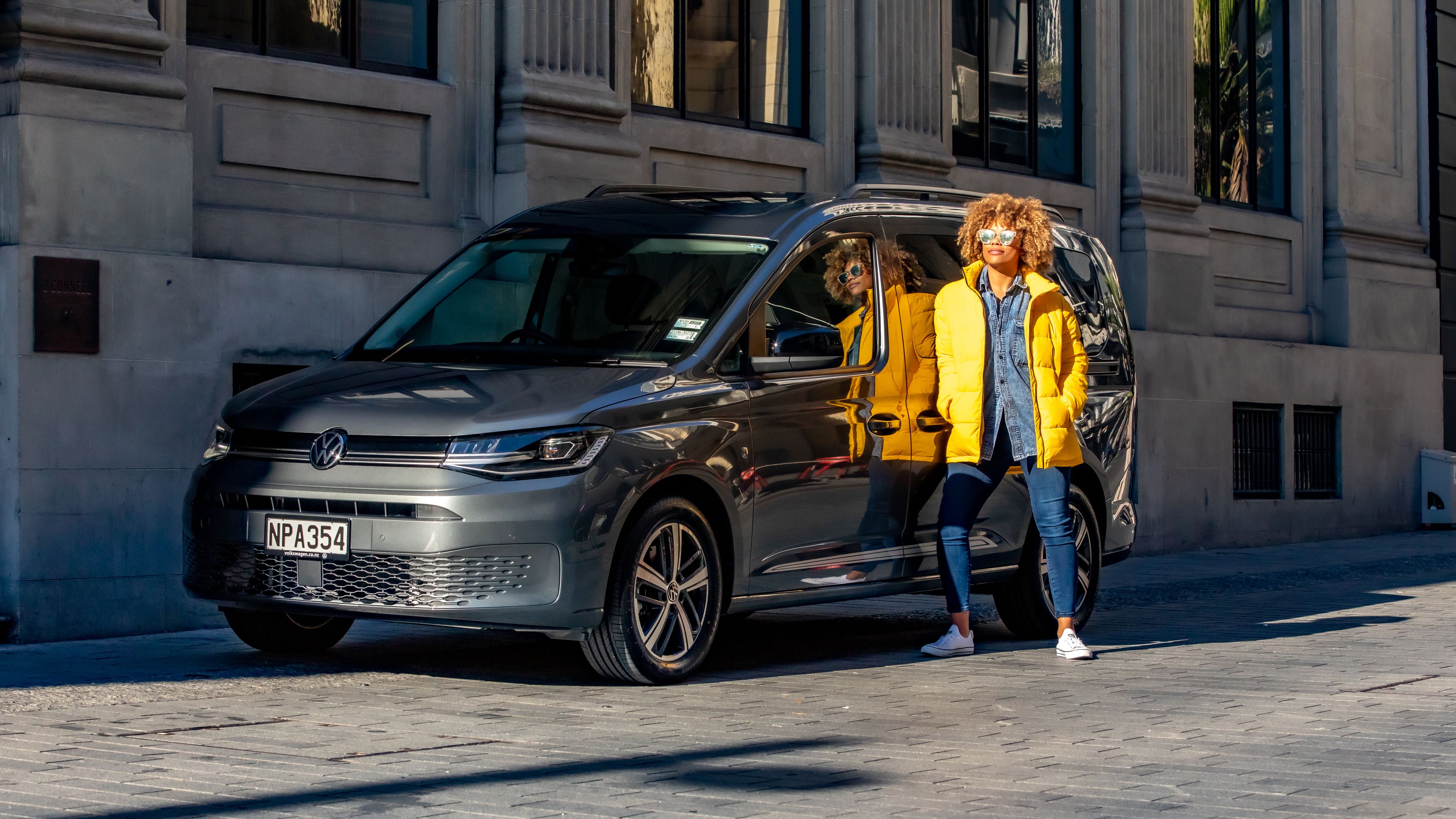Oseba z rdečkastimi kodrastimi lasmi v svetlo rumeni puhovki samozavestno stoji naslonjena na srebrn enoprostorec Volkswagen, parkiran ob mestni ulici. Popoldansko sonce ustvarja močan odsev na vratih avtomobila in poudari barvo jakne. V ozadju so visoke kamnite stavbe z okni in pilastri, ki dajejo prizoru urbano, evropsko vzdušje.